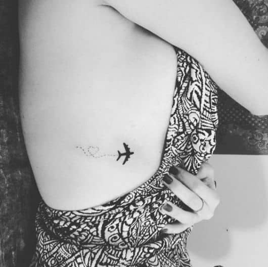 Tatuagem delicada do contorno de um avião preenchido de preto que deixa um rastro em forma de coração enquanto voa. 