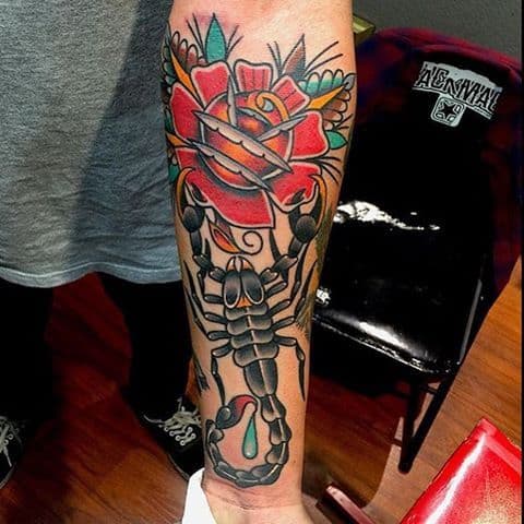 Tatuagem colorida no antebraço de um escorpião e uma rosa. Na ponta do ferrão dele está saindo uma gota de veneno. 
