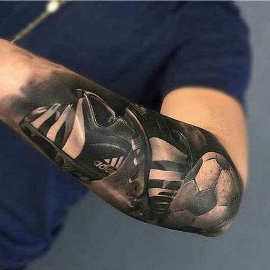Tatuagem no antebraço de um par de chuteiras da adidas ao lado de uma bola de futebol. 