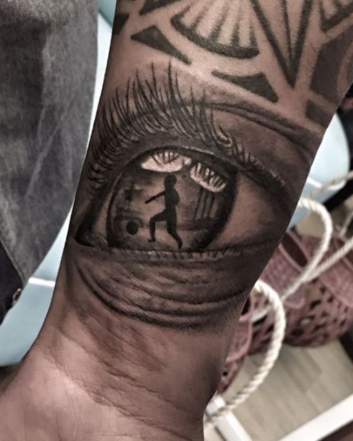 Tatuagem próxima ao pulso com o desenho de um olho realista e em suas pupilas é possível observar um garoto jogando futebol. 
