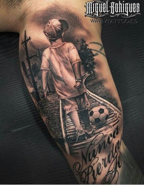 Tatuagem na parte interna do braço de um menino andando no trilho de trem com uma bola de futebol em seus pés. 