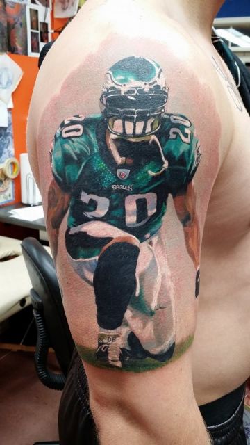 Tatuagem colorida no braço com o desrnho de um jogador de futebol americano de joelhos. 