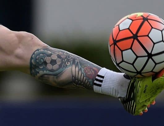 Perna com uma chuteira e meião de futebol chutando uma bola. A panturrilha é coberta por uma tatuagem futebol. 