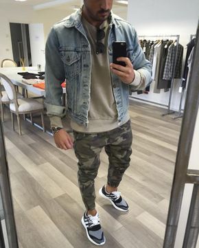 Homem tirando uma selfie em uma loja de roupa. Ele veste uma jaqueta jeans sobreposta a um moletom liso e uma calça swag apertada com estampa militar. 