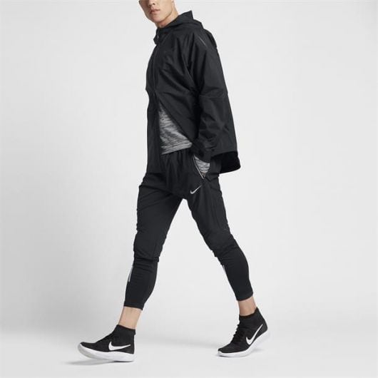 Homem em um fundo branco caminhando enquanto veste uma calça justa da Nike. Ele também veste uma jaqueta preta fechada e tênis da Nike. 