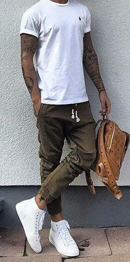 Foto do corpo de um homem tatuado segurando uma mochila enquanto encosta na parede. Ele veste uma calça swag com elástico no calcanhar e com caimento não tão baixo. 