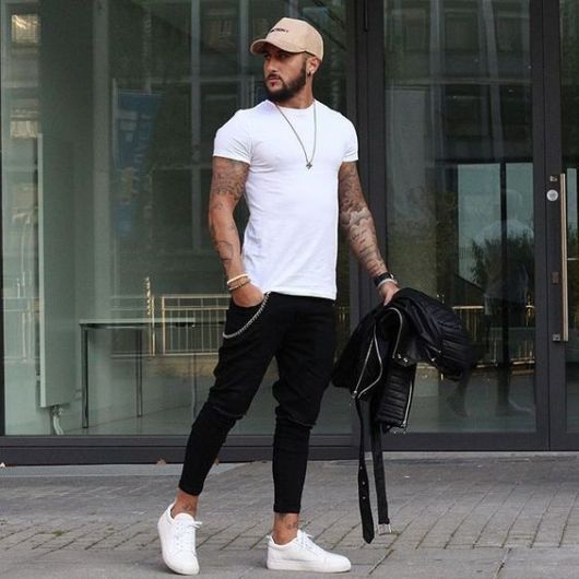 Homem de boné e camiseta lisa simples caminhando enquanto segura uma jaqueta de couro. Ele veste uma calça swag jeans skinny. 