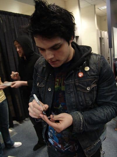 O cantor Gerard Way segurando uma caneta prestando atenção no que está escrevendo. Ele veste um moletom com estampa de super-heróis e uma jaqueta com dois botons. 