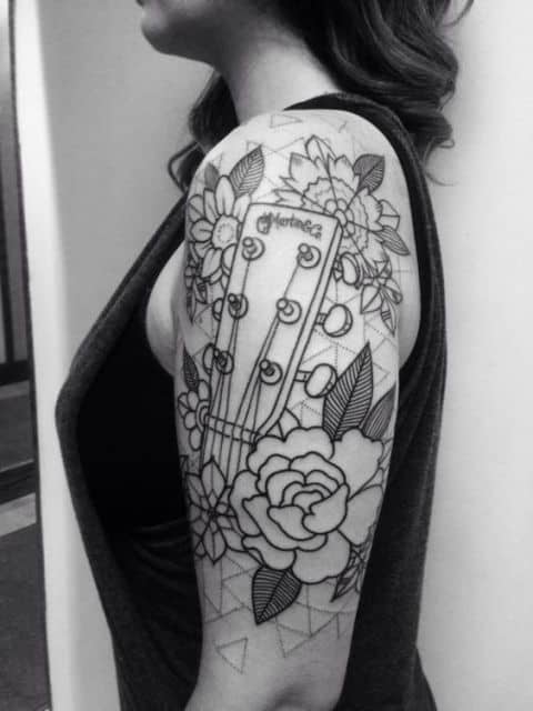 Tatuagem na parte superior do braço de uma mulher com a 'mão' de una guitarra e diversas flores ao seu redor. 