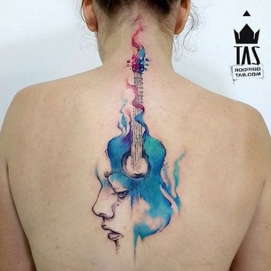 Tatuagem no centro das costas de uma mulher com um violão feito com aquarela e um rosto saindo de uma das laterais de sua base. 