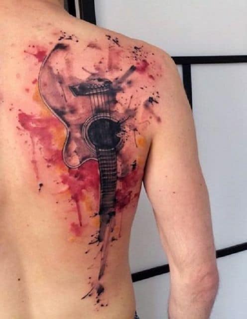 Tatuagem grande nas costas de um homem com um violão se desfazendo e cores vivas de aquarela ao seu redor. 