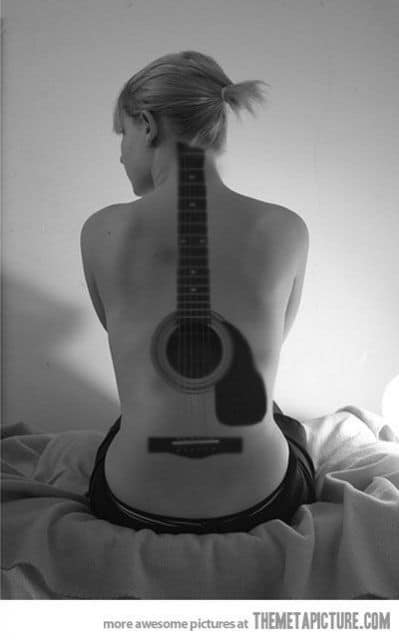 Tatuagem grande que vai do pescoço até a parte baixa das costas de uma mulher. Não há o contorno do violão, apenas o braço, as cordas, a boca e o rastilho. 