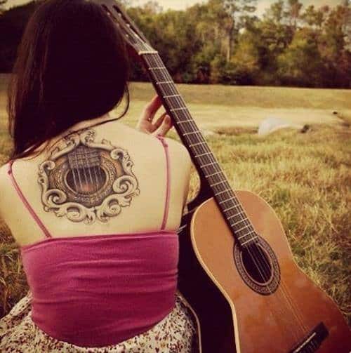 Tatuagem redonda no centro das costas de uma mulher acompanhada de seu violão. A tatuagem apenas revela a boca e parte das cordas do violão. 