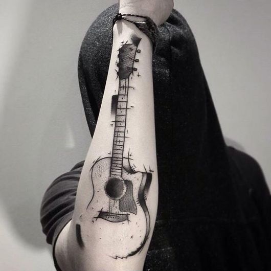 Homem com o rosto coberto e o braço levantado, onde há uma tatuagem de violão feita como se fosse um esboço na parte externa do antebraço. 