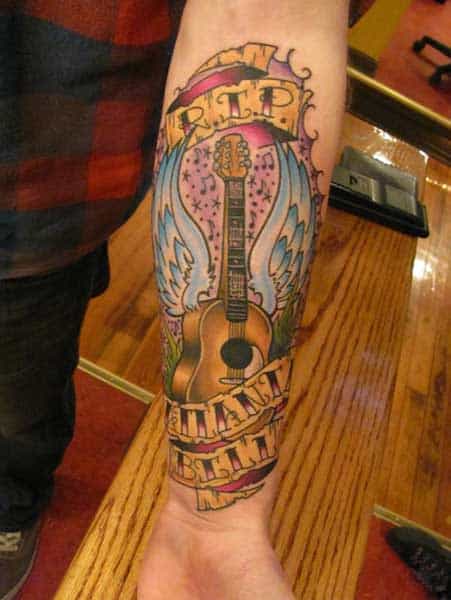 Tatuagem colorida de uma violão com asas e uma faixa que faz homenagem a alguém quem faleceu. 