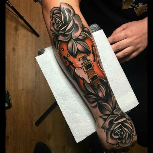 Tatuagem na parte externa do antebraço com o corpo de uma guitarra e flores ao seu redor. 