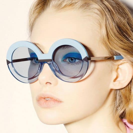Modelo de cabelos presos usa óculos de sol azul com armação azul bebe.