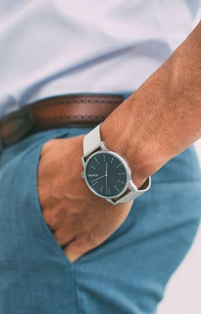Foto do pulso de um homem com a mão no bolso. Ele usa um relógio prata arredondado sem muitos detalhes, apenas ponteiros e os números que indicam as horas. 