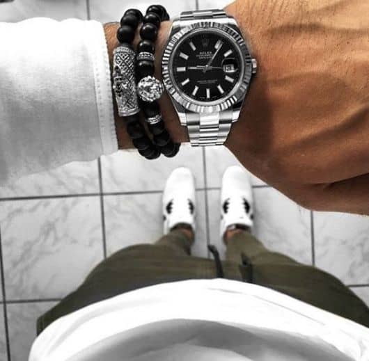 Foto do pulso de um homem com trajes casuais com foco no relógio bem elaborado acompanhado de duas pulseiras. 