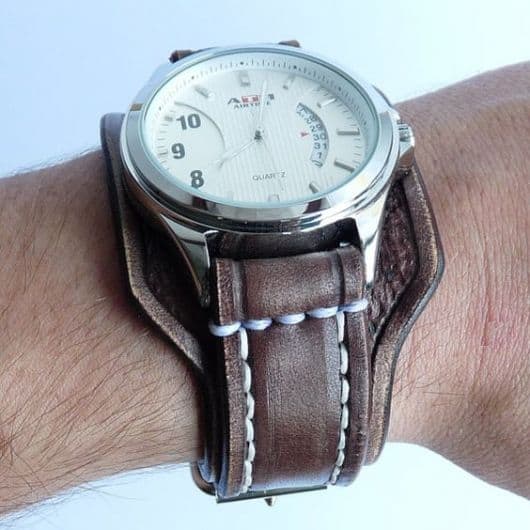 Foto de um relógio com aro prata arredondado e uma pulseira grossa de couro. 