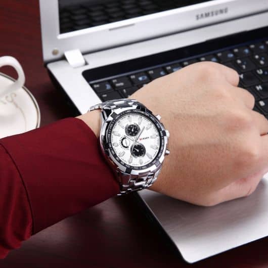 Foto do pulso de um homem posicionado em frente a um notebook. O relógio conta com diversos detalhes e funcionalidades, além de um design ousado e chamativo. 