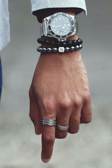 Foto do pulso de um homem usando duas pulseiras e dois anéis. Acima das pulseiras está um relógio prata muito detalhado no aro e na pulseira. 