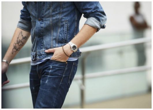 Foto do corpo de um homem caminhando na rua vestindo jaqueta e calça jeans. Em um de seus pulsos está um relógio prata em formato arredondado. 