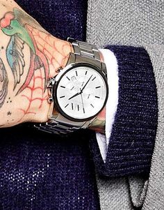 Foto do pulso e parte da mão de um homem vestindo roupas de frio. Acima de sua mão tatuado está um relógio prata chique com ponteiros minimalistas. 