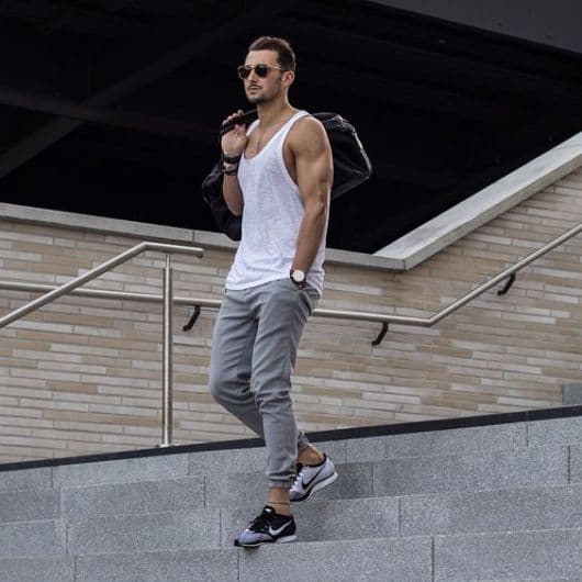 Homem descendo uma escada. Ele veste uma camiseta regada cavada, calça moletom jogger, tênis da Nike, óculos escuros e um relógio prata arredondado. 