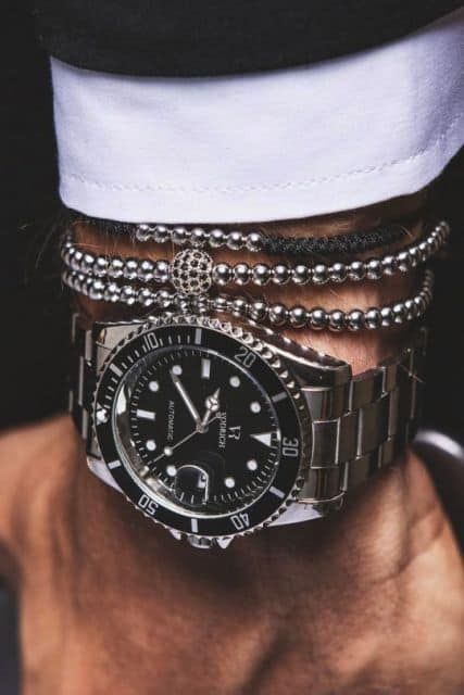 Foto do pulso de um homem com parte de um traje social acompanhado por pulseiras e um relógio prata repleto de detalhes. 