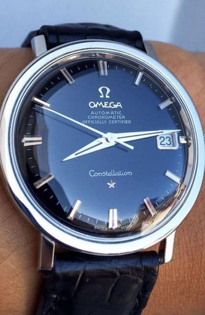 Foto de um relógio da marca Omega arredondado com pulseira de couro e pulseira prata. 