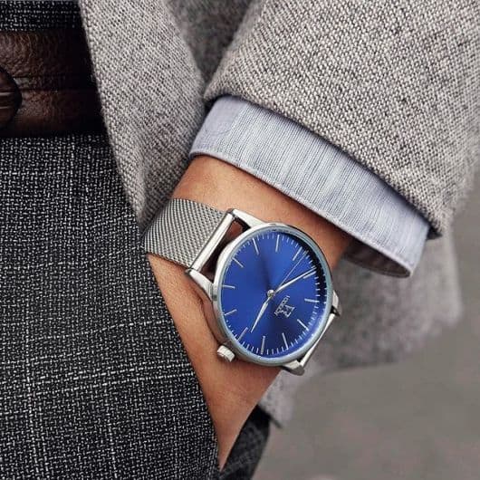 Foto do pulso de um homem com a mão no bolso vestindo um traje social. Ele usa um relógio arredondado prata com poucos botões ou detalhes. O destaque fica na pulseira que tem uma textura em relevo. 
