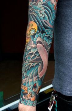 Tatuagem cobrindo o braço com traços orientais. Vemos o fundo do mar com um tubarão ao lado de um mergulhador. 