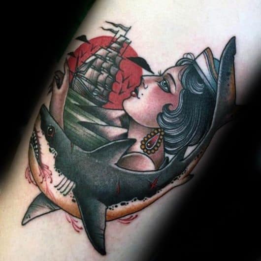 Tatuagem no estilo tradicional de um tubarão com sangue saindo de sua boca e cicatrizes em seu corpo. Acima dele há uma mulher de perfil e uma caravela com o sol vermelho ao fundo. 