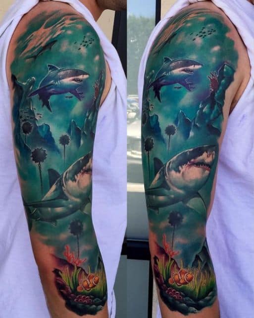 Tatuagem cobrindo o braço de um homem. Há o fundo do mar muito detalhado com dois tubarões brancos nadando. 