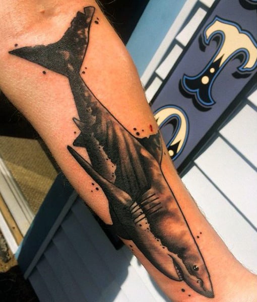 Tatuagem de tubarão no antebraço. Ele é feito com tons de cinza e é visto de perfil enquanto nada.