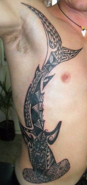 Tatuagem grande na costela de um homem. Há um tubarão martelo que sai de seu ombro e vai até sua cintura feito no estilo maori. 