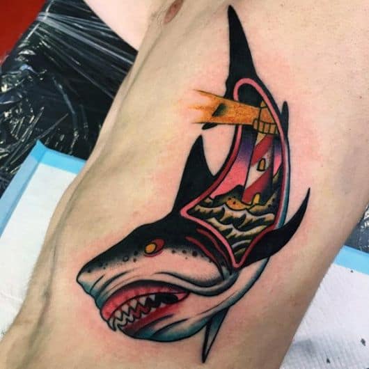 Tatuagem na costela seguindo o estilo Old School com um tubarão agressivo e uma abertura no interior de seu corpo onde há um farol em águas agitadas. 