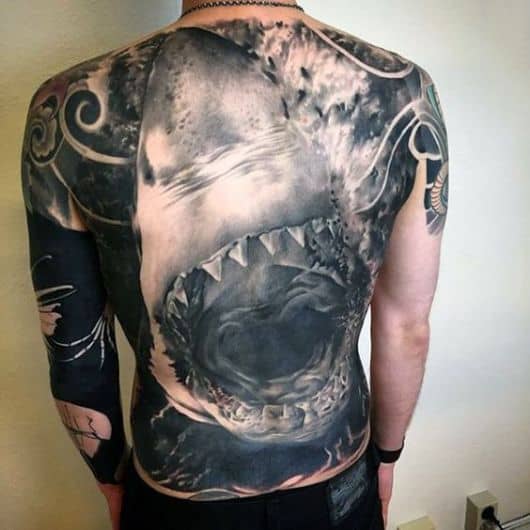 Tatuagem realista de um tubarão saindo da água com as mandíbulas abertas em posição de ataque cobrindo as costas inteiras de um homem.