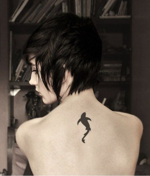 Tatuagem pequena da silhueta de um tubarão feita no centro das costas de uma mulher. 