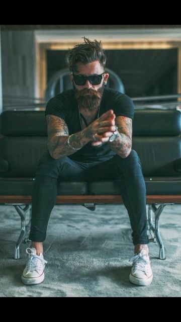 Homem tatuado com trajes simples e escuros sentado em um sofá. Ele usa um óculos quadrado grande com lentes e hastes escuras. 