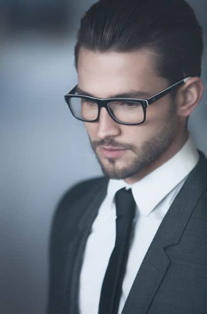 Homem de terno, gravata e cabelo penteado para trás usando um óculos quadrado de grau com hastes pretas e pequenos detalhes em branco. 