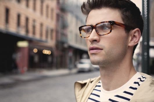 Foto de um homem na rua em primeiro plano com o fundo desfocado. Ele usa um óculos quadrado de grau cujas hastes misturam diferentes tipos de cor. 
