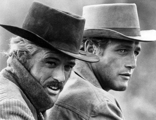 Paul Newman e Robert Redford usando trajes típicos de cowboys americanos, inclusive o chapéu. 