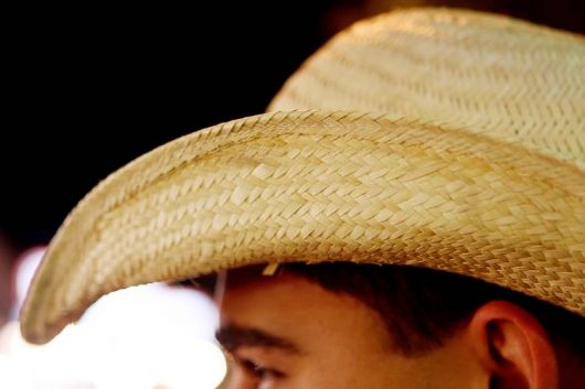 Foto com foco em um chapéu de palha na cabeça de um homem. A palha é bem entrelaça para não deixar espaços em branco no chapéu. 