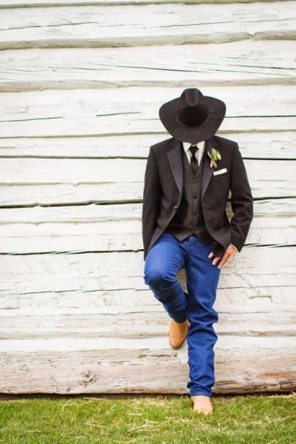 Homem recostado na parede de madeira. Ele usa bota, calça jeans, terno, colete, gravata e um chapéu country masculino preto. 