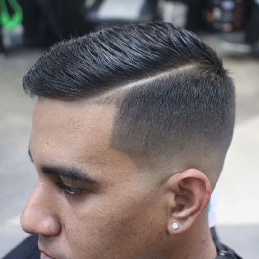Homem de perfil na barbearia com corte militar penteado para o lado no topo e lateral bem raspada com um risco na parte superior. 