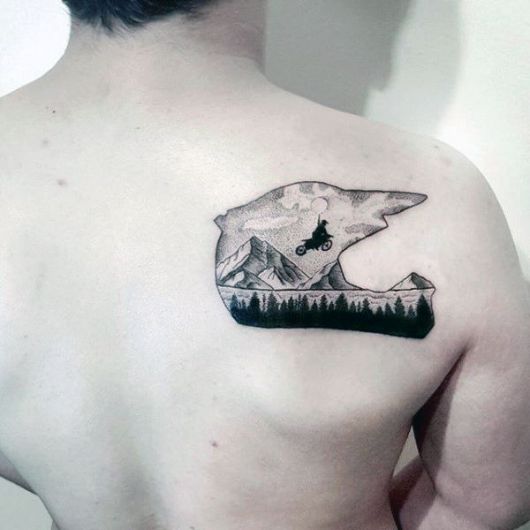 Tatuagem nas costas de um homem com o formato de um capacete e dentro dele uma paisagem onde um motoqueiro está pulando alto com sua moto. 