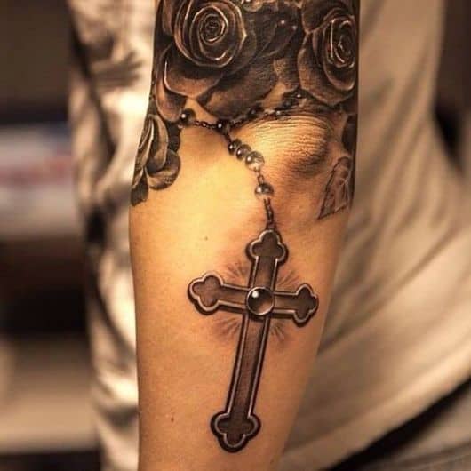 Tatuagem em tons de cinza próxima ao cotovelo de uma mulher com o desenho de uma cruz bem detalhada. 