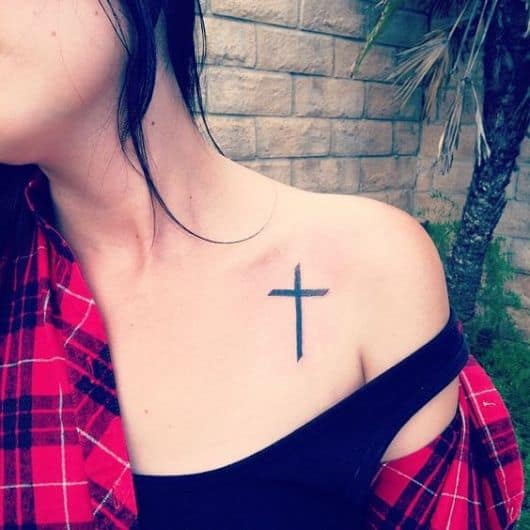 Tatuagem religiosa delicada na parte frontal do ombro de uma mulher com o desenho de uma cruz pintada de preto. 